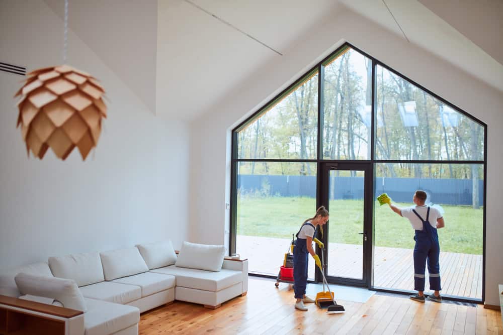 Two people in uniforms from Deep Cleaning DuPage clean a modern living room with high ceilings and large windows. One person sweeps the hardwood floor while the other wipes the glass door. The room features a white sectional sofa and a wooden light fixture, with trees visible outside.