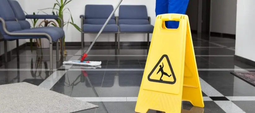 A person mops the floor in a hallway with blue chairs. A yellow caution sign featuring a slipping figure is prominently displayed on the shiny tiled surface to warn of the wet floor. Nearby, plants and mats complete the scene, ensuring thorough Office Cleaning DuPage County style.