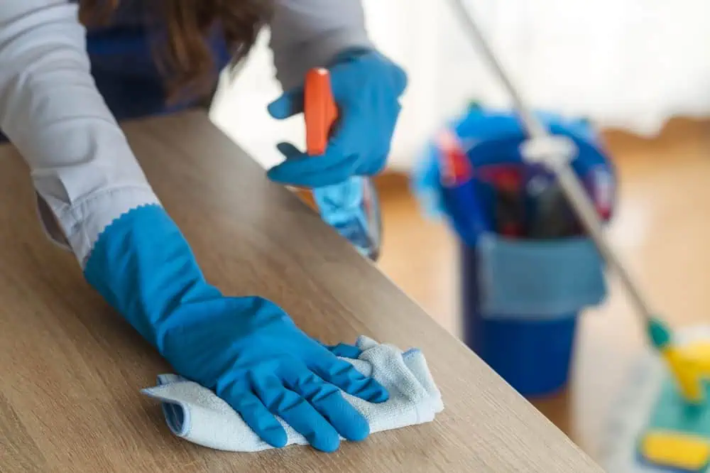 A person wearing blue gloves cleans a wooden surface with a cloth while holding a spray bottle. A blue cleaning bucket with various supplies and a mop, characteristic of Residential Cleaning DuPage County services, are visible in the background.