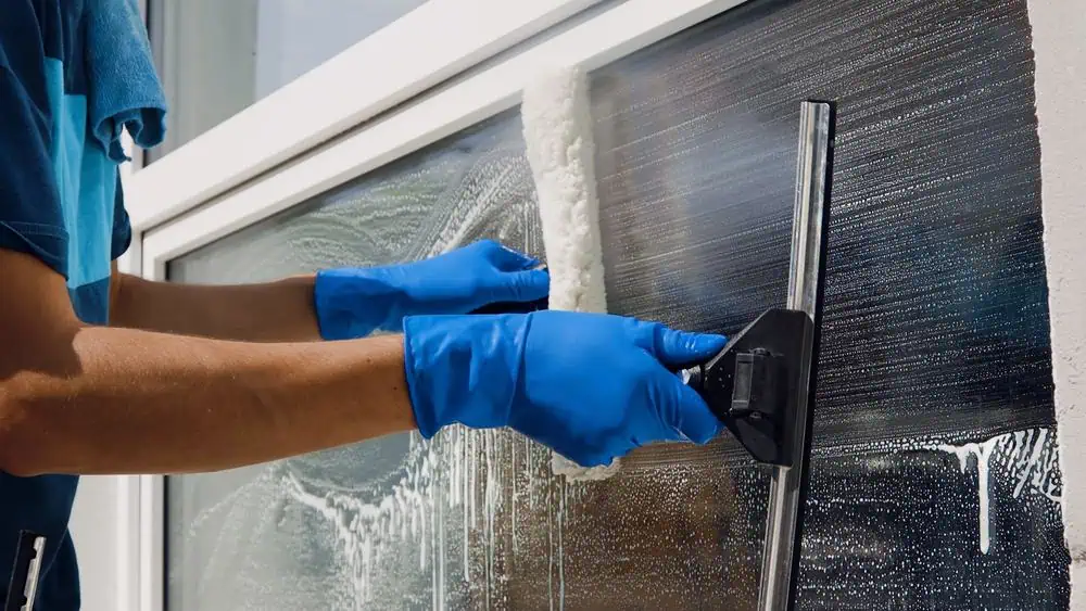 A person wearing blue gloves cleans a window with a squeegee. The window, partially covered with soap suds, indicates that it's being washed. The person's upper body and the window frame are visible, demonstrating thorough attention to detail typical of deep cleaning in DuPage County.