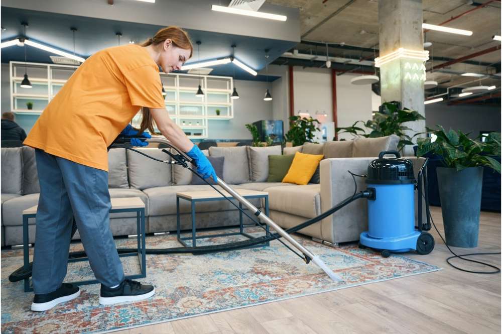 A person wearing an orange shirt and blue gloves uses a carpet cleaning machine on a colorful rug in a modern, open-concept room with a gray couch, plants, and contemporary lighting—showcasing top-notch residential cleaning in DuPage County.