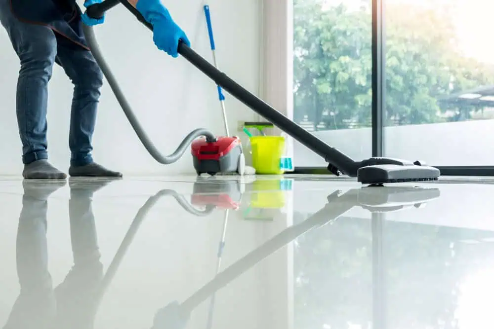 A person wearing blue gloves is vacuuming a shiny, tiled floor with a vacuum cleaner. Cleaning supplies, including a yellow bucket and mop, are in the background next to large windows overlooking a green outdoor area. This scene exemplifies the meticulous attention to detail of Commercial Cleaning DuPage County services.