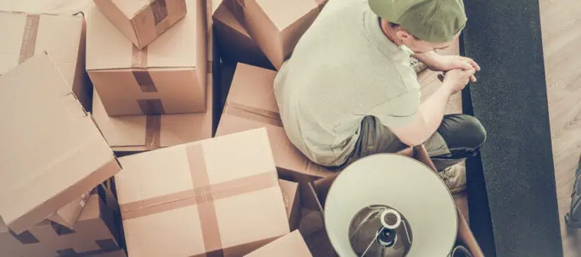 A person wearing a cap sits amidst a pile of cardboard boxes and rolled-up carpet, suggesting a moving process. The individual appears to be taking a break and looking at something in their hands. A lampshade is visible on one of the boxes, likely awaiting Deep Cleaning DuPage Cleaning services.