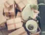 A person wearing a cap sits amidst a pile of cardboard boxes and rolled-up carpet, suggesting a moving process. The individual appears to be taking a break and looking at something in their hands. A lampshade is visible on one of the boxes, likely awaiting Deep Cleaning DuPage Cleaning services.