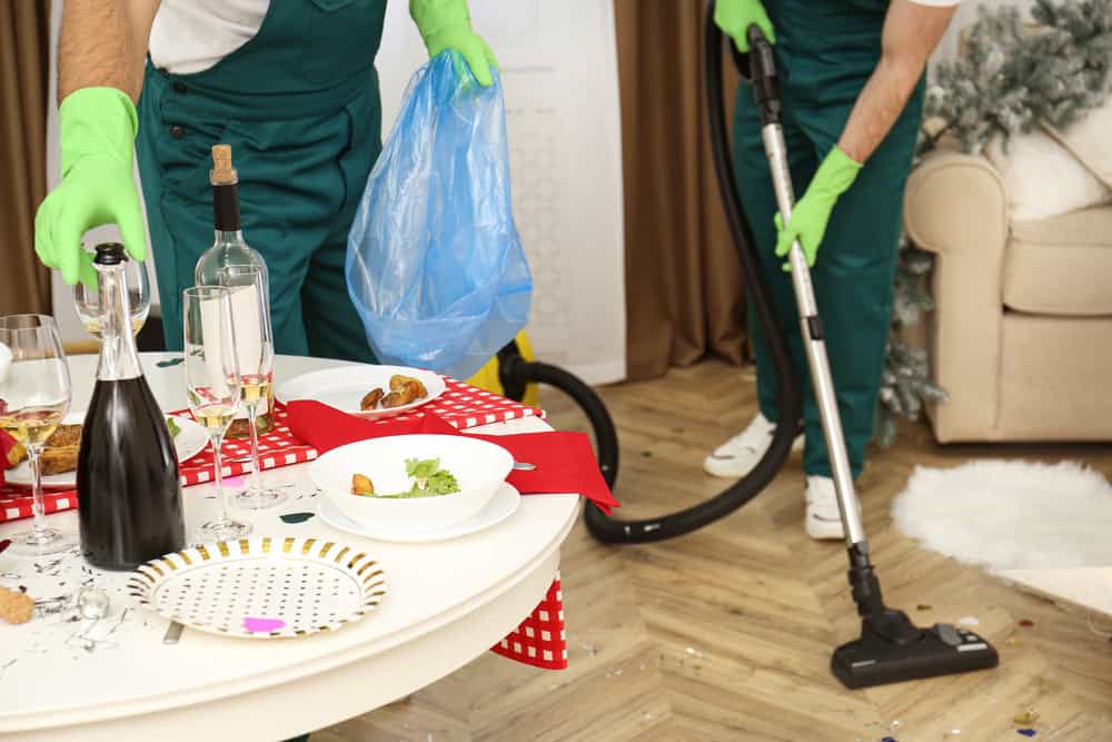 Two people in green overalls and gloves are performing a deep cleaning in DuPage County. One is vacuuming the floor while the other holds a blue trash bag near a table with leftover food and drinks. The table is cluttered with dirty dishes, and the floor is littered with confetti.