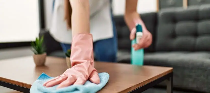 A person wearing pink cleaning gloves wipes a wooden coffee table with a blue cloth in a living room, showcasing the meticulous attention to detail synonymous with Residential Cleaning DuPage County. They hold a spray bottle in the other hand, while a small potted plant adorns the table and a gray sofa sits in the background.