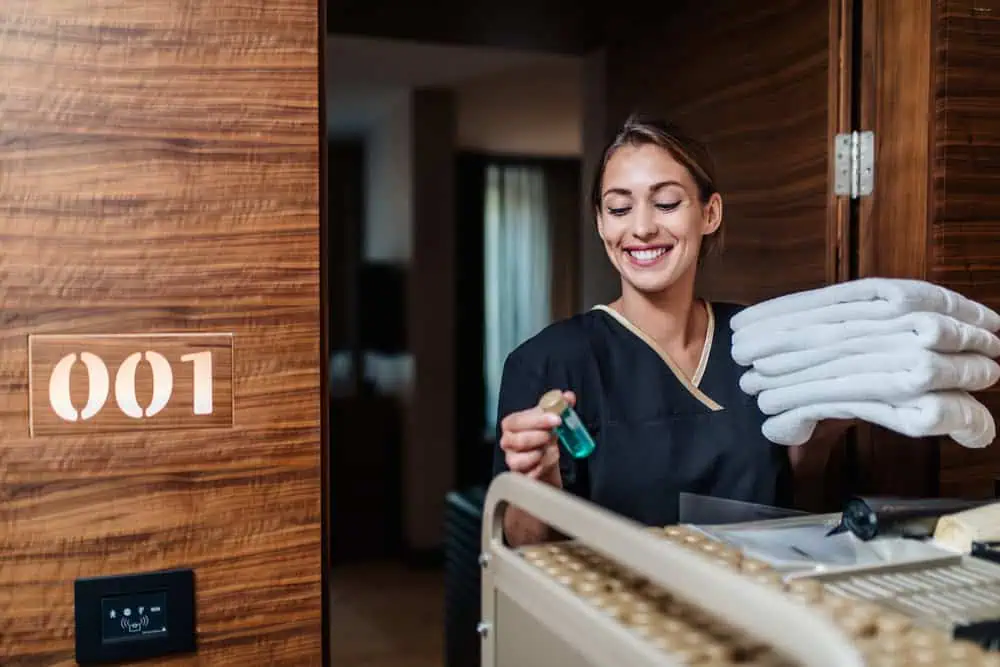 A housekeeping staff member in uniform holds a small bottle of cleaning solution with several folded towels stacked on her arm. She stands next to a wooden door labeled "001," appearing to prepare for office cleaning in DuPage County. She is smiling warmly, ready to restock the room.