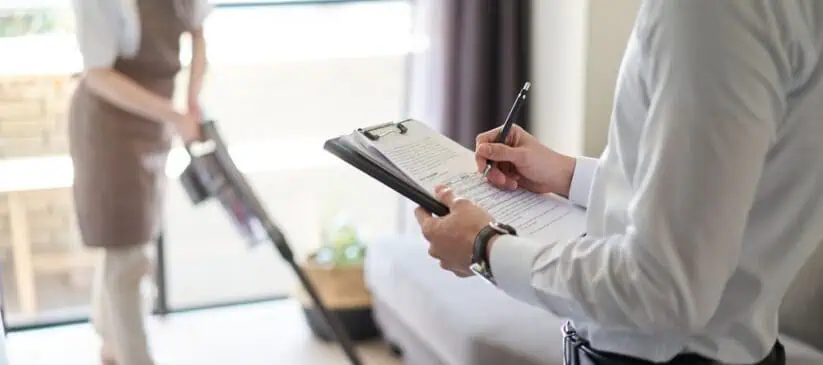 A person in a white shirt holding a clipboard and pen observes someone in the background wearing an apron and vacuuming the carpet, showcasing the meticulous attention to detail provided by Deep Cleaning DuPage County in a living room with a sofa and large window.