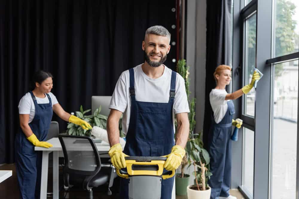 Three workers, wearing uniforms and gloves, are cleaning an office space. The foreground features a man smiling while operating a cleaning machine. In the background, one person is cleaning a desk area while another cleans a large window—exemplifying top-notch Commercial Cleaning DuPage County services.