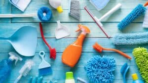 Various cleaning supplies including brushes, scrubbers, sponges, a spray bottle, dustpans, and gloves arranged on a blue wooden surface. The items are colorful with shades of blue, green, orange, red, and yellow—ideal for any Residential Cleaning DuPage County tasks.