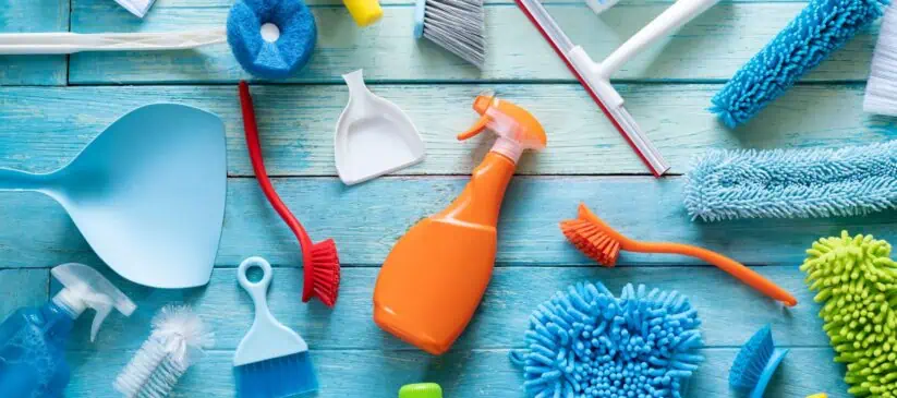 Various cleaning supplies including brushes, scrubbers, sponges, a spray bottle, dustpans, and gloves arranged on a blue wooden surface. The items are colorful with shades of blue, green, orange, red, and yellow—ideal for any Residential Cleaning DuPage County tasks.