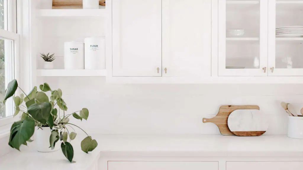 A bright, minimalist kitchen features white cabinets and shelves. A potted plant sits on the countertop next to containers labeled "Sugar" and "Flour." Cutting boards and a marble slab lean against the backsplash. Light shines through a window on the left, showcasing a space ready for deep cleaning DuPage.