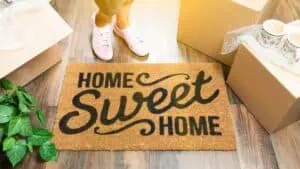 A person wearing pink sneakers stands on a wooden floor next to a doormat that reads "Home Sweet Home" in black text. Surrounding the mat are cardboard boxes and a potted plant, suggesting a new home scene. Residential Cleaning DuPage County can help keep your space spotless as you settle in.