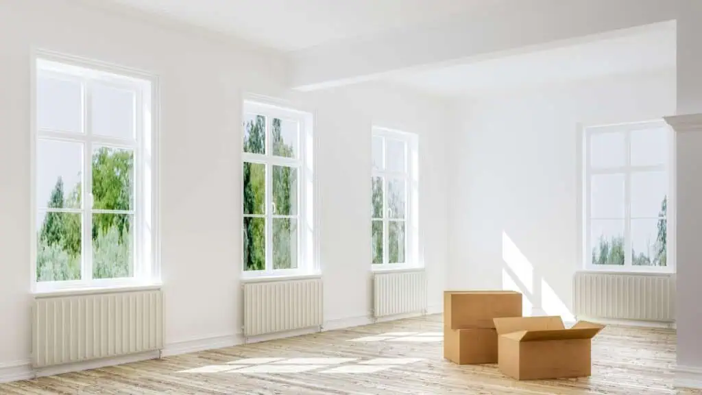 A bright, unfurnished room with large windows, white walls, and wooden floors. Natural light streams into the room, and two brown cardboard boxes are on the floor, suggesting a moving or unpacking scene. Perfect for Deep Cleaning DuPage Cleaning services to help you start fresh. The view outside includes lush green trees.