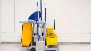 A janitor's cart against a plain white wall. The cart is equipped with a large yellow bag, several cleaning tools including brooms, mops, and dusters, a yellow mop bucket, cleaning solutions, and various other janitorial supplies essential for deep cleaning DuPage County offices.