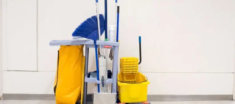 A janitor's cart against a plain white wall. The cart is equipped with a large yellow bag, several cleaning tools including brooms, mops, and dusters, a yellow mop bucket, cleaning solutions, and various other janitorial supplies essential for deep cleaning DuPage County offices.