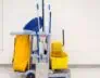 A janitor's cart against a plain white wall. The cart is equipped with a large yellow bag, several cleaning tools including brooms, mops, and dusters, a yellow mop bucket, cleaning solutions, and various other janitorial supplies essential for deep cleaning DuPage County offices.