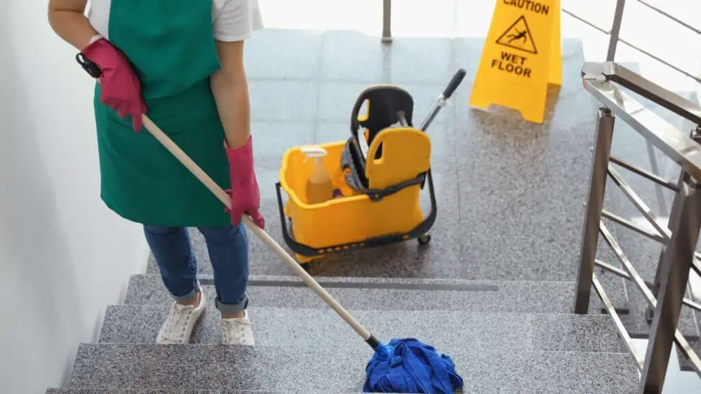 A person in a green apron and pink gloves is mopping a set of stairs. A yellow cleaning bucket with a wringer and a mop handle is on the landing. A yellow "Caution Wet Floor" sign is in the background near a metal railing, illustrating top-notch Office Cleaning DuPage County services in action.