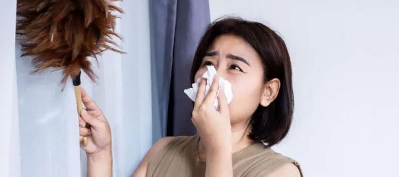 A woman with short dark hair is holding a feather duster in one hand while covering her nose with a tissue, possibly due to allergies. A curtain hangs in the background. For thorough relief and a dust-free home, consider Deep Cleaning DuPage County services for top-notch residential cleaning needs.