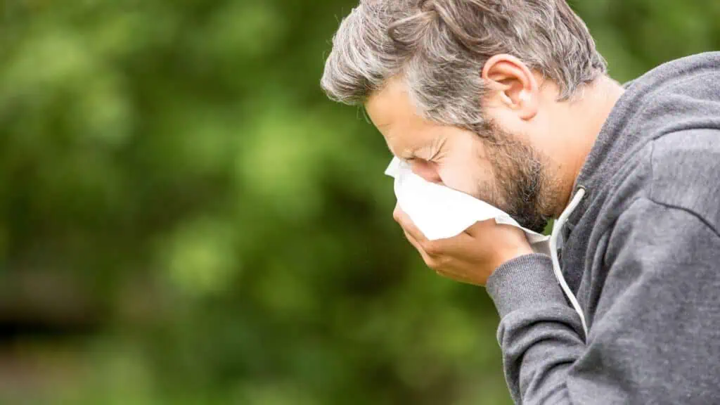 A man with gray hair and a beard is outdoors in a green, blurry setting, sneezing into a tissue. He is wearing a dark hoodie, and his eyes are closed as he holds the tissue to his face, possibly thinking about the Residential Cleaning DuPage County services he recently hired.