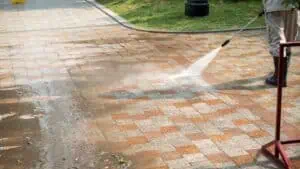 A person is power washing a stone-tiled patio with a high-pressure hose, visibly cleaning and removing dirt, revealing a clear distinction between the cleaned and uncleaned areas. Grass is in the background, and the worker in boots and trousers efficiently handles this Commercial Cleaning DuPage County task.