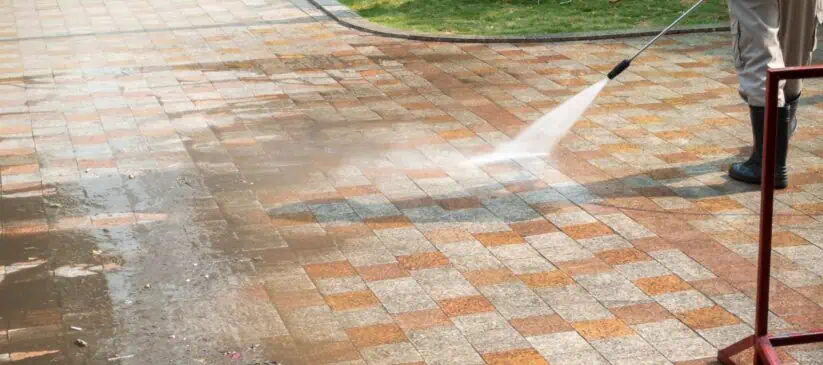 A person is power washing a stone-tiled patio with a high-pressure hose, visibly cleaning and removing dirt, revealing a clear distinction between the cleaned and uncleaned areas. Grass is in the background, and the worker in boots and trousers efficiently handles this Commercial Cleaning DuPage County task.