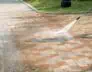 A person is power washing a stone-tiled patio with a high-pressure hose, visibly cleaning and removing dirt, revealing a clear distinction between the cleaned and uncleaned areas. Grass is in the background, and the worker in boots and trousers efficiently handles this Commercial Cleaning DuPage County task.