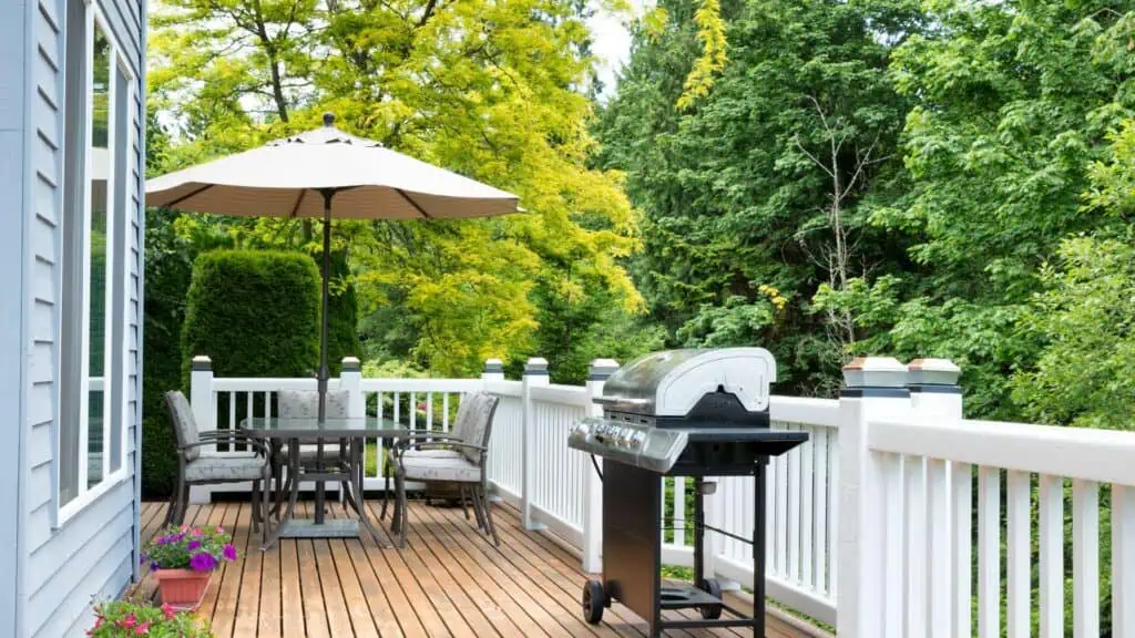 A wooden deck featuring a black grill and a patio set with a white umbrella is shown. The patio set includes a table and four chairs. The deck, perfect for relaxation after Office Cleaning DuPage County, is bordered by white railings and surrounded by lush green trees. A small potted plant with pink flowers is placed in the corner.