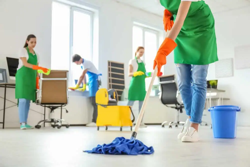 A group of four people wearing green aprons and orange gloves clean an office. One person is mopping the floor in the foreground, while others are tidying desks and cleaning windows in the background. A blue bucket and cleaning supplies are visible.