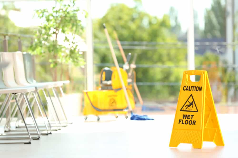 A yellow "Caution Wet Floor" sign is placed on a shiny white floor in a bright, spacious room with large windows. In the background are several chairs, a mop, and a bucket on a cleaning cart. Sunlight filters through the windows, illuminating the space.