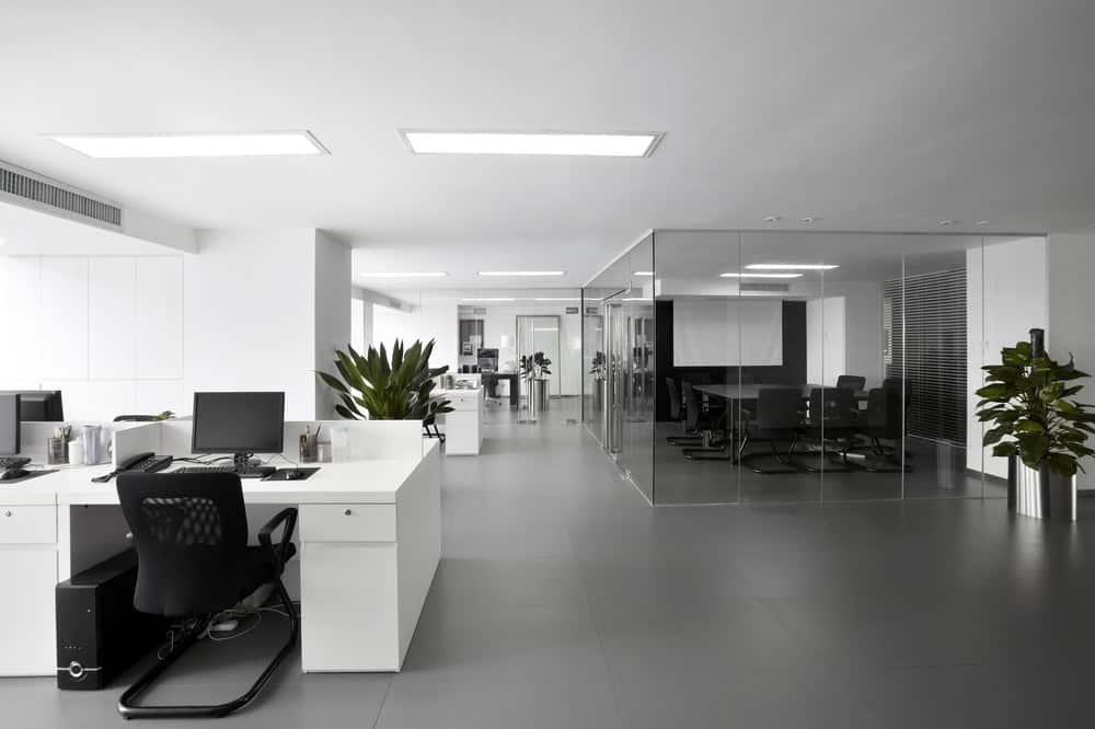 A modern, minimalistic office space featuring white desks with computer monitors, ergonomic chairs, and green potted plants. A glass-walled conference room is in the background, and the floor is covered with sleek, gray tiles. Bright ceiling lights illuminate the area. Ensure your workspace always shines with top-notch Office Cleaning DuPage County services.