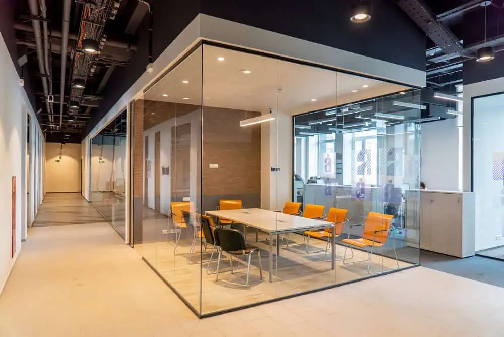 A modern office meeting room with glass walls, adorned with orange chairs around a wooden table. The room is well-lit with ceiling lights. Adjacent corridors are visible through the glass, showcasing an open office layout with various workspaces.