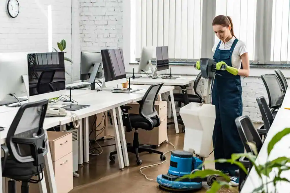 A person in overalls and gloves is cleaning the floor with a machine in a modern office space with multiple workstations, computers, and office chairs. The office, benefiting from Commercial Cleaning DuPage County services, features large windows with blinds and potted plants visible in the foreground.