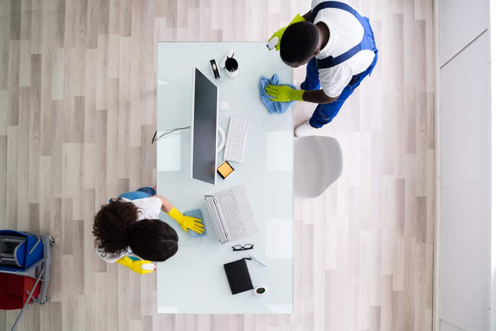 Two people are cleaning an office workspace—one wipes a desktop while the other, in blue coveralls, cleans a glass-top table. Items on the desk include a computer, keyboard, mouse, notebook, smartphone, and coffee mug. Nearby is a cleaning cart from Office Cleaning DuPage County services.