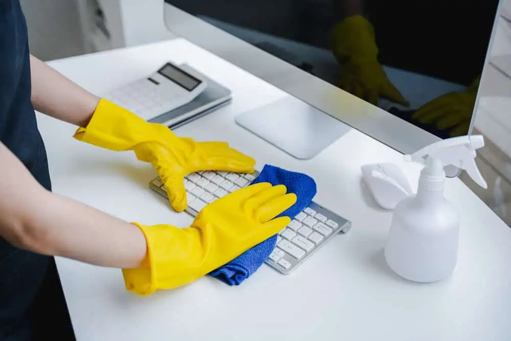 A person wearing yellow gloves is meticulously cleaning a computer keyboard with a blue cloth. On the desk, there's also a bottle of cleaning solution and a calculator, exemplifying the thoroughness of deep cleaning in DuPage County.