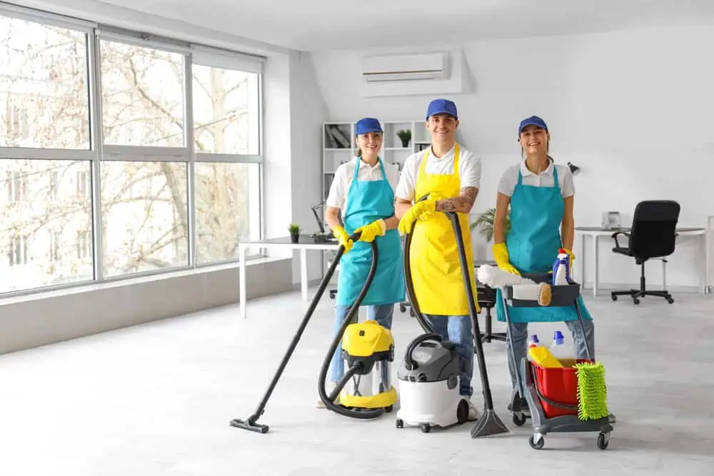 Three smiling cleaning staff members pose in a bright, modern office. They wear uniforms, caps, gloves, and aprons (yellow and blue). Cleaning tools include a vacuum, mop, and supply cart. Large windows and a tidy desk are visible in the background.