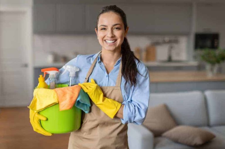 House cleaning service professional holding a bucket with cleaning supplies, gloves, and cloths inside a modern living room and kitchen area.